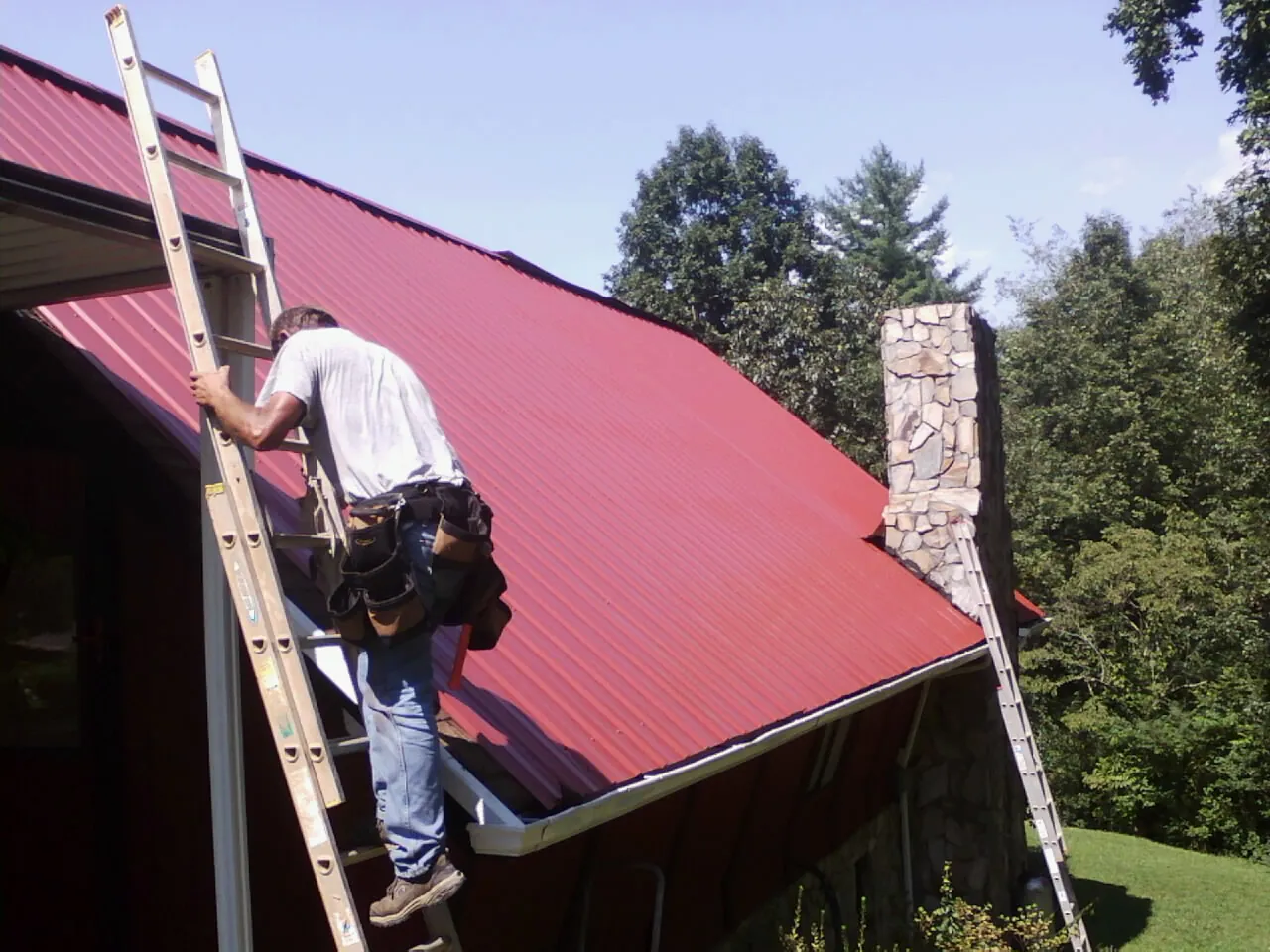 Roofer on a ladder beside a red metal roof and stone chimney — Soffit & Fascia Repair by Roofing Brothers in Lane City